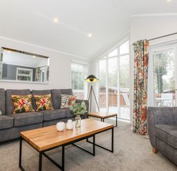 A living room with a gray sofa and armchair a wooden coffee table and side table a floor lamp and large windows with floral curtains at Squirrels Nook Waterside Wood 19 White Cross Bay near Windermere