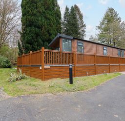 A brown wooden cabin with a fenced deck surrounded by trees at Squirrels Nook in Waterside Wood 19 White Cross Bay near Windermere