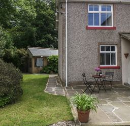 An outdoor patio area with two chairs and a table with flowers and a bottle next to a house and a small shed in the garden at Gardeners Cottage in Millom Cumbria