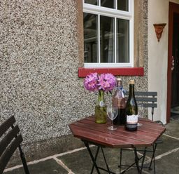 A small outdoor table with two chairs beside a pebble wall with a window a vase with flowers and two bottles on the table at Gardeners Cottage in Millom Cumbria