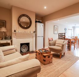 A living room with a sofa, armchair, wooden chest table, white fireplace, round mirror, and a dining area with a table and chairs at 3 Albion Cottages in Walberswick