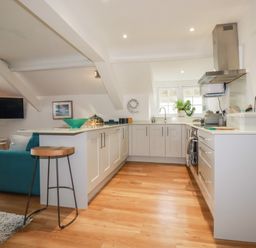 A kitchen with white cabinets and wooden floor connected to a living area with a teal sofa at Harbour Light in Boscastle