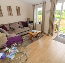 A living room with a beige sofa and purple cushions a small wooden coffee table a glass side table with books and a lamp wooden floor and sliding glass doors leading outside at Garden View in Pembroke