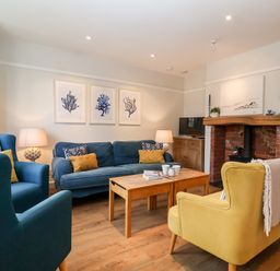 A living room with a blue sofa, yellow armchair, wooden coffee table, fireplace, TV, and framed art on the wall at Fawcett Cottage in Aldeburgh