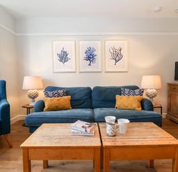 A living room with blue sofa and armchair yellow and patterned cushions wooden coffee tables and framed artworks at Fawcett Cottage in Aldeburgh