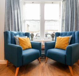 Two blue armchairs with yellow and patterned cushions in front of a window with grey curtains at Fawcett Cottage in Aldeburgh