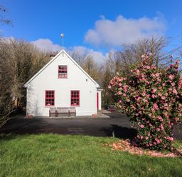 A white house with red window frames and benches outside surrounded by trees and a flowering bush in grass at Kyleatunna Cottage in Ennis County Clare