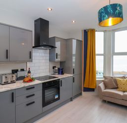 A kitchen with grey cabinets and appliances next to a beige sofa with pillows by windows with yellow curtains at Apt 6 @ Hunter's Quay in Bridlington