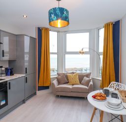A kitchen and dining area with gray cabinets, a small sofa near windows with yellow curtains, and a table set with croissants and dishes at Apt 6 @ Hunter's Quay in Bridlington