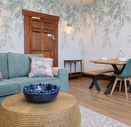 A living room with a green sofa and patterned cushions a round gold coffee table and a wooden dining table with green chairs at Watermouth Castle West Tower Apartment in Berrynarbor near Ilfracombe