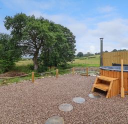 An outdoor gravel area with a wooden hot tub and steps near trees and fields at Heron Pod in Nantmawr near Trefonen