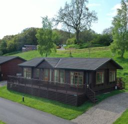 A wooden cabin with a deck and a gravel driveway surrounded by green grass and trees at The Fairhaven in Tunstall near Catterick North Yorkshire