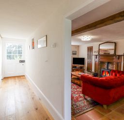 A hallway with wooden flooring leads to a white door and opens into a living room with a red sofa a wooden coffee table a fireplace and a television at West House Farm in Leiston