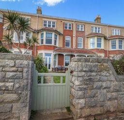 A stone wall with a green wooden gate leading to a garden and a row of brick terraced houses at Blue Yonder in Weymouth