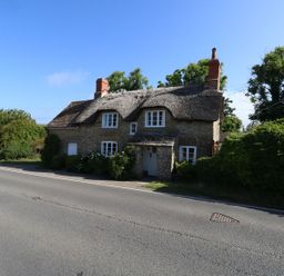 A stone house with a thatched roof and two chimneys next to a road with greenery around Alice's Cottage Burton Bradstock