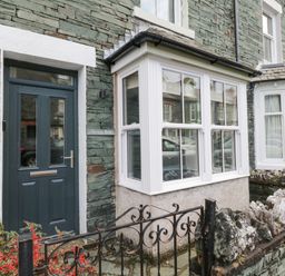 The exterior of a stone house with a dark door and white framed bay windows behind a black metal gate with red berries and rock wall at Forty Five in Keswick