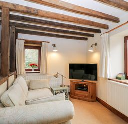 A small living room with a patterned sofa wooden beams on ceiling a TV on wooden stand and flowers on window sills at Well Cottage in Moreton-In-Marsh