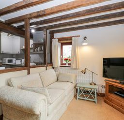 A living room with a patterned sofa a small side table with a lamp a window with curtains and a vase of flowers and a television on a wooden stand at Well Cottage in Moreton-In-Marsh
