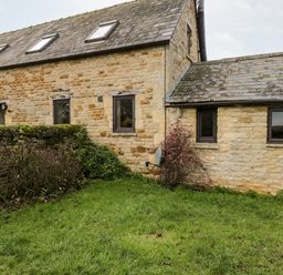 An exterior view of a stone house with multiple windows a stone wall and grass at Stable Cottage in Moreton-In-Marsh