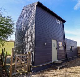 An exterior view of a dark grey metal sided building with a small beige door and steps surrounded by a wooden gate and gravel at The Stables in Tafarn-Y-Gelyn