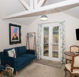 A room with a blue sofa a small round table with two chairs a wall-mounted TV and glass doors with curtains at The Potting Shed in Combe near Langport