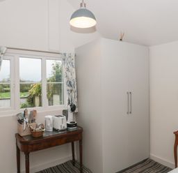 A room with a window with floral curtains a wooden table with kitchen appliances a white wardrobe and a small wooden side table at The Potting Shed in Combe near Langport