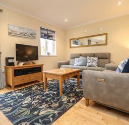 A living room with grey sofas a wooden coffee table and TV stand a patterned blue rug and wall art at Manuka in Falmouth