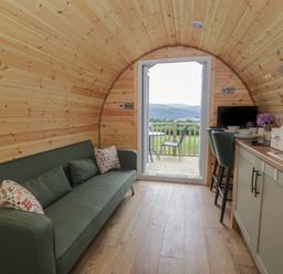 A pod interior with a green sofa, kitchen area with stools, and an open door to a deck with chairs at Tarrenhendre Pod in Llanegryn near Tywyn
