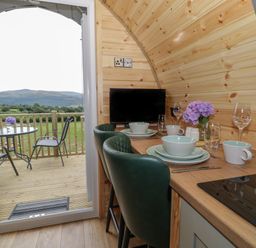 A small dining area with plates, bowls, cups, wine glasses, a television, and flowers on a wooden counter with green chairs and an open door leading to a balcony with a table and chairs in the countryside at Tarrenhendre Pod in Llanegryn near Tywyn