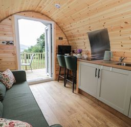 A small wooden pod interior with a green sofa pillows kitchen area with sink kettle two chairs and an open door to a deck with outdoor chair at Tarrenhendre Pod Llanegryn near Tywyn