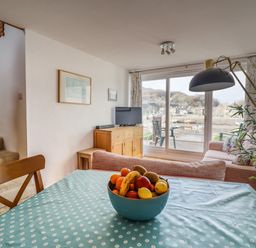 A dining table with a bowl of fruit in a living room with sofas a television and sliding glass doors overlooking a harbor at Branwen Porthmadog