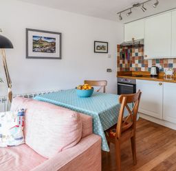 A kitchen with a dining table covered with a blue tablecloth and chairs next to a pink sofa with a cushion and a floor lamp in Branwen Porthmadog