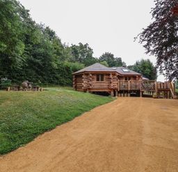 A log cabin with a wooden deck and stairs beside a dirt path with a grassy area and outdoor fireplace at Copper Lodge in Chulmleigh