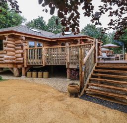 A wooden log cabin with a raised deck and stairs surrounded by trees at Copper Lodge in Chulmleigh