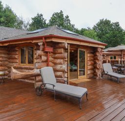 An outdoor wooden deck with two cushioned lounge chairs a wooden log cabin and a dining table with an umbrella at Copper Lodge in Chulmleigh