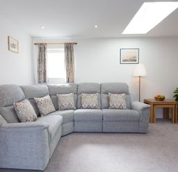 A living room with a gray sectional sofa with patterned pillows a floor lamp wooden nesting tables with a fruit bowl and a potted plant at Cornish Views in Camborne