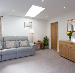 A living room with a grey sofa and patterned pillows next to wooden tables and a wooden cabinet with a mirror at Cornish Views in Camborne