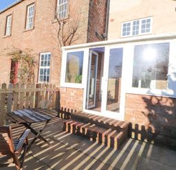 An outdoor patio with wooden table and chairs next to a brick house with a white framed glass door at Hillside Hideaway in Wigton