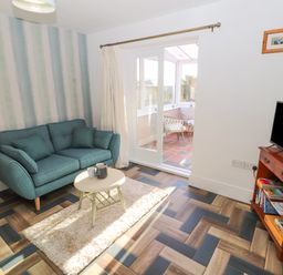 A living room with a teal sofa a small round table a television on a wooden stand and sliding glass doors leading to a sunroom at Hillside Hideaway in Wigton