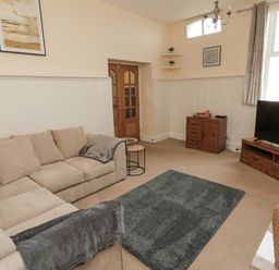 A living room with a beige sectional sofa a gray rug a wooden TV stand with a television a wooden cabinet corner shelves and framed pictures at Courtyard Cottage in Alnwick
