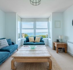 A living room with two blue sofas a wooden coffee table a side table with a lamp and a window with a sea view at Tamarama House in Port Isaac