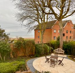 An outdoor seating area with a round paved patio table and chairs near trees and shrubs by red brick buildings at 9 Church Farm Rise in Aldeburgh