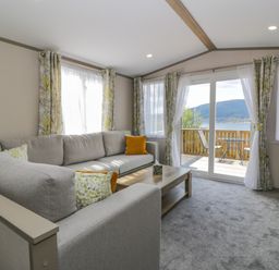 A living room with a gray sectional sofa and cushions by windows and glass doors opening to a balcony with chairs and a table at 11A Mansion View in Cove near Kilcreggan