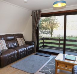 A living room with a brown leather sofa, two plaid cushions, a wooden coffee table with books, rugs on the floor, glass sliding doors, and a view of a wooden balcony and trees at 27 Invergarry Lodges in South Laggan near Invergarry