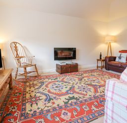 A living room with a patterned rug a wooden rocking chair a leather sofa a plaid armchair and a wall-mounted fireplace at Wellington Lodge Cottage in Middleton Tyas