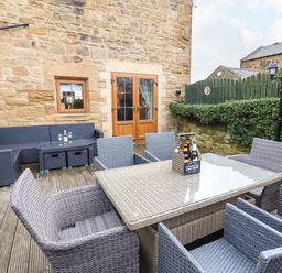 An outdoor patio with a wicker dining table and chairs and a wicker sofa set against a stone building at Winding Wheel Cottage in Cambois near Ashington