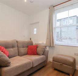 A small living room with a beige sofa with red cushions and a matching ottoman near a window with blinds and curtains at Jacks Cottage in Keswick