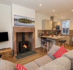 A living room with a fireplace a wall-mounted television a kitchen island with two stools and a sofa with red cushions at Jack's Cottage in Keswick
