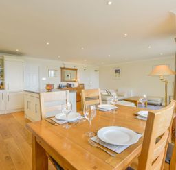 An open kitchen and dining area with a wooden table set for four and a living area with a sofa and floor lamp at 19 Burgh Island Causeway in Bigbury-On-Sea
