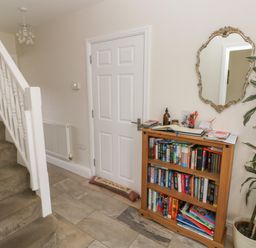 A hallway with carpeted stairs a wooden bookshelf with books a potted plant and a wall mirror at Llygad Yr Haul Sun in Burry Port